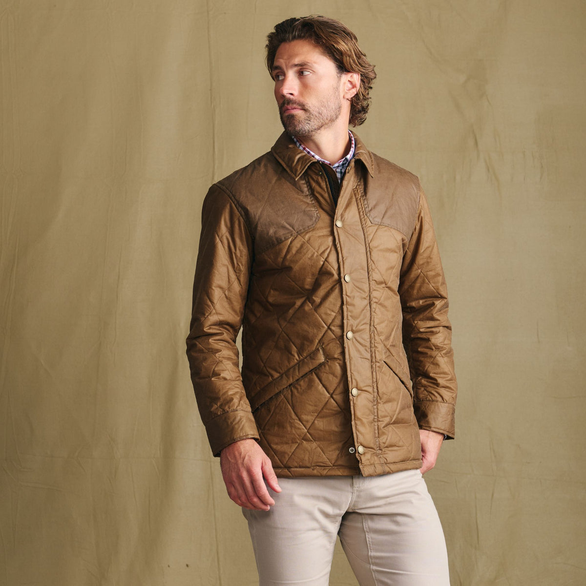 Man in a Tom Beckbe Paddock Quilted Jacket and light pants stands against a tan background, looking left.