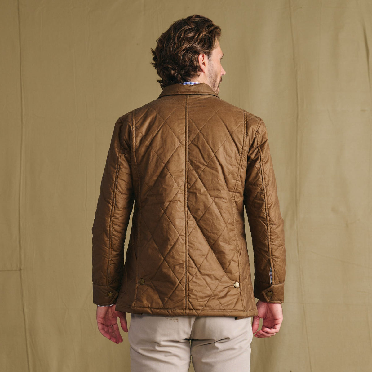 Man facing away in Tom Beckbe Paddock Quilted Jacket and light pants, standing before a plain tan background.