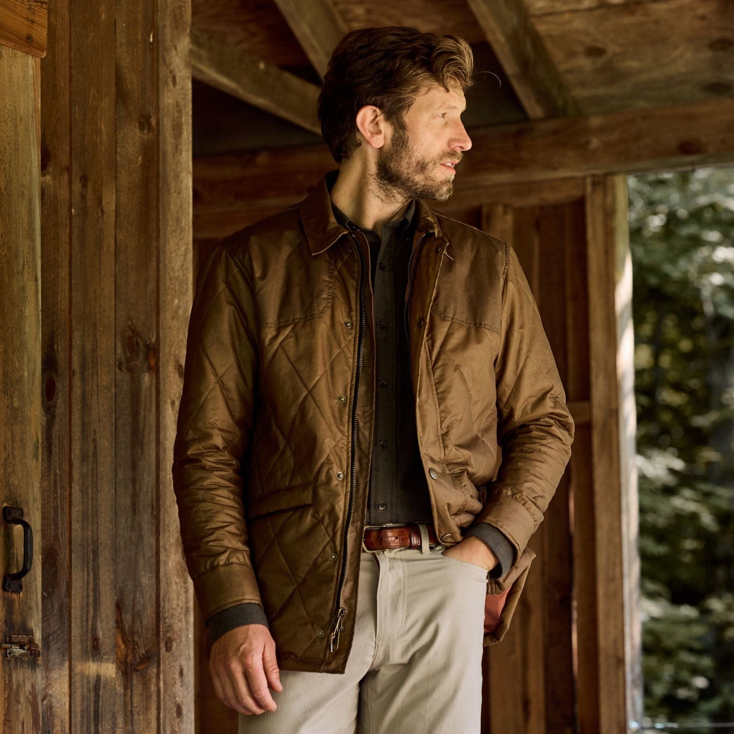A man in a Tom Beckbe Paddock Quilted Jacket and light pants stands on a porch, glancing sideways with trees behind.