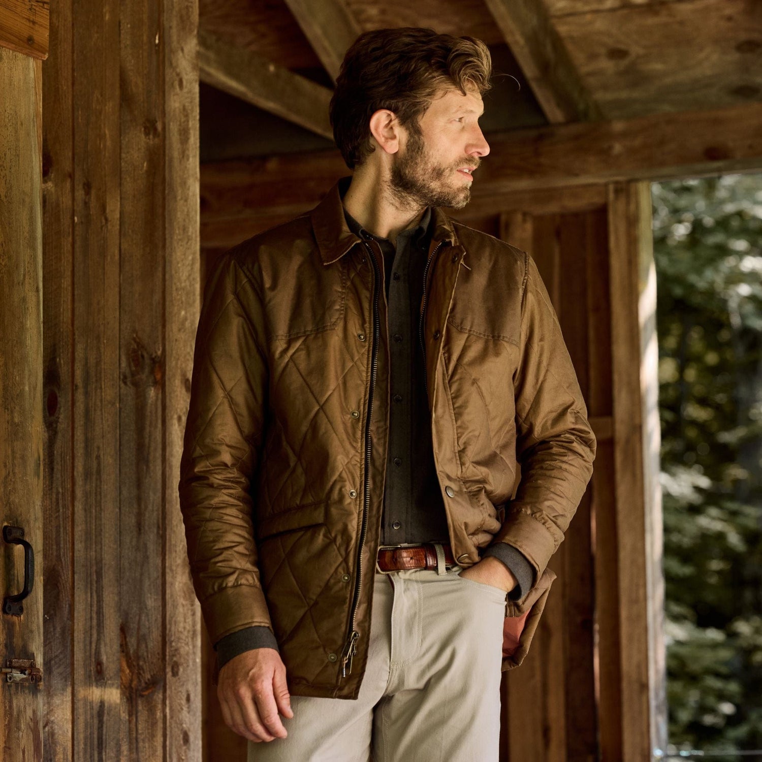 A man in a Tom Beckbe Paddock Quilted Jacket and light pants stands on a porch, glancing sideways with trees behind.