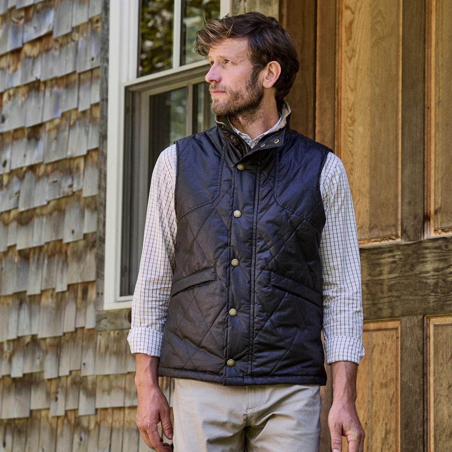 A man in a Tom Beckbe Paddock Quilted Vest and checkered shirt stands by a wooden house, looking left.