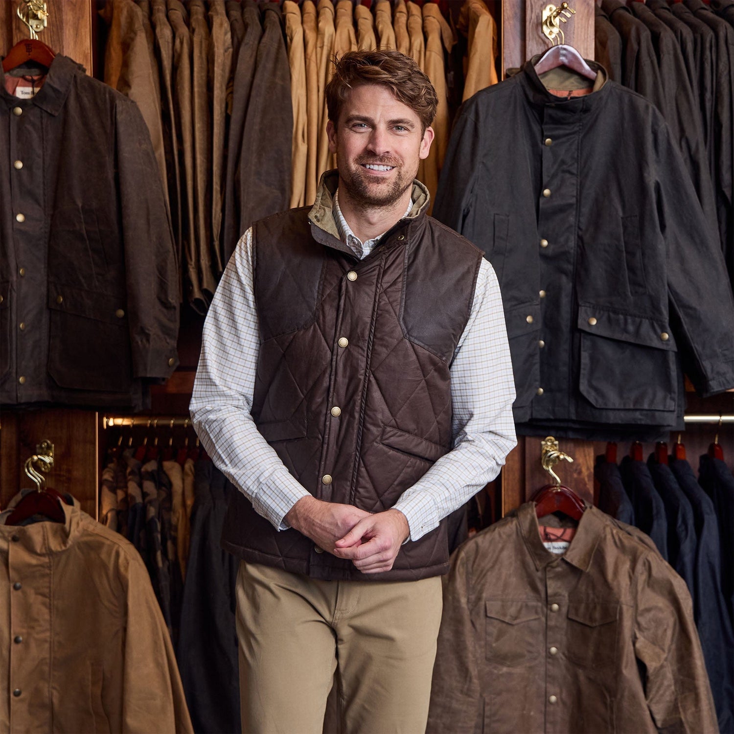 Man wears Tom Beckbe Paddock Quilted Vest over a checked shirt and khaki pants, standing by a jacket display.