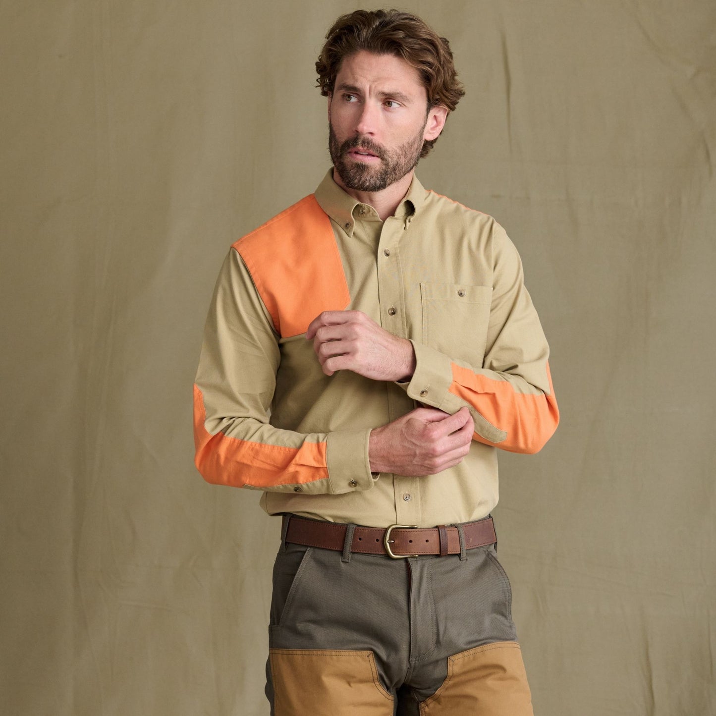 Man in Tom Beckbe Perry Field Shirt with orange accents, color-blocked pants, stands before a plain tan backdrop.