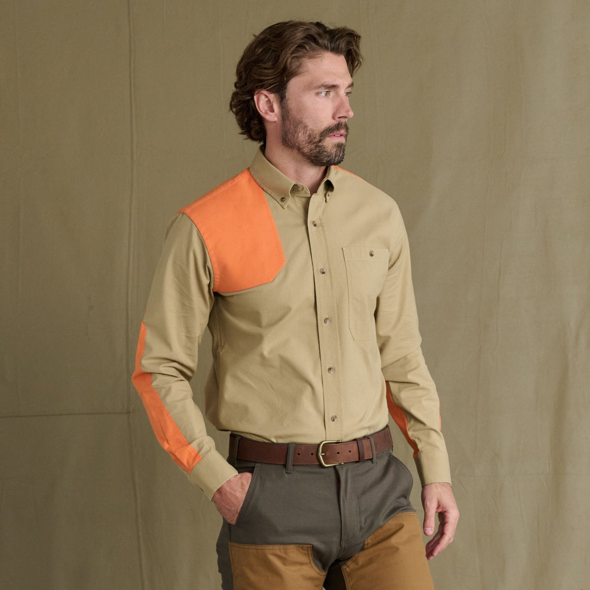 A man wears the Tom Beckbe Perry Field Shirt in beige with orange accents and two-tone brown pants, facing left.