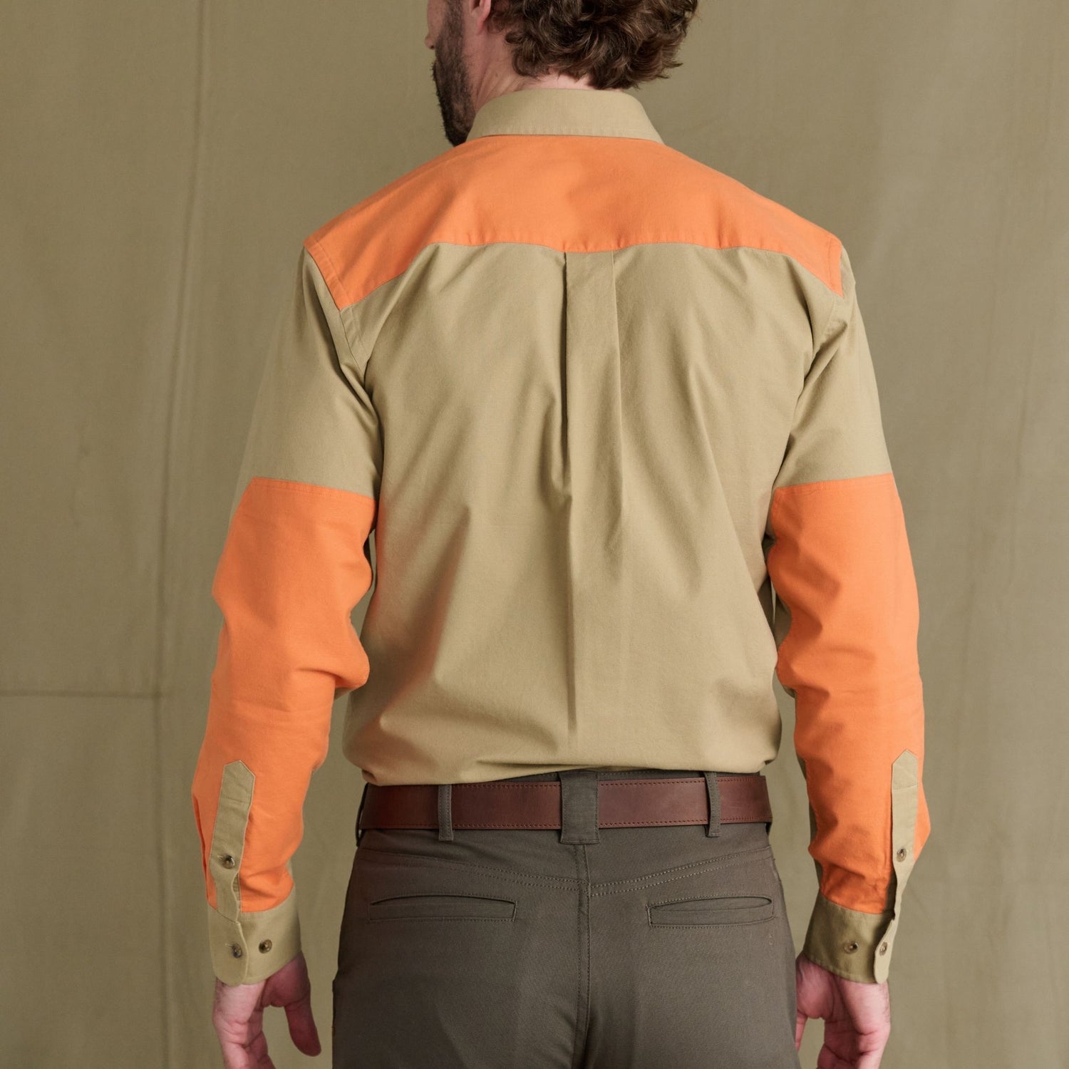 Man facing away in Tom Beckbe Perry Field Shirt with orange shoulders, sleeves, and olive pants; plain background.