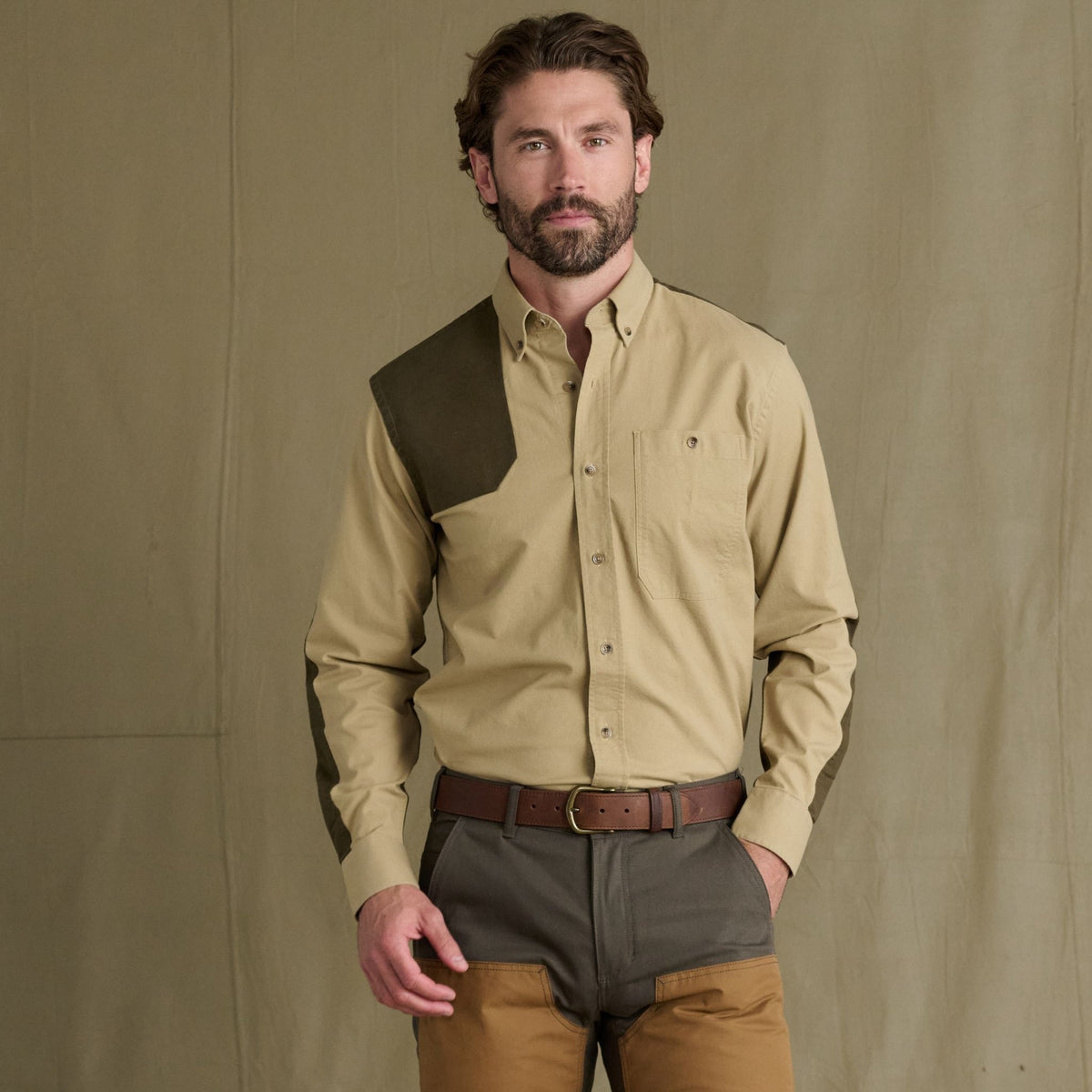 Man wearing Tom Beckbe Perry Field Shirt with green shoulder patch and brown pants, against a beige background.