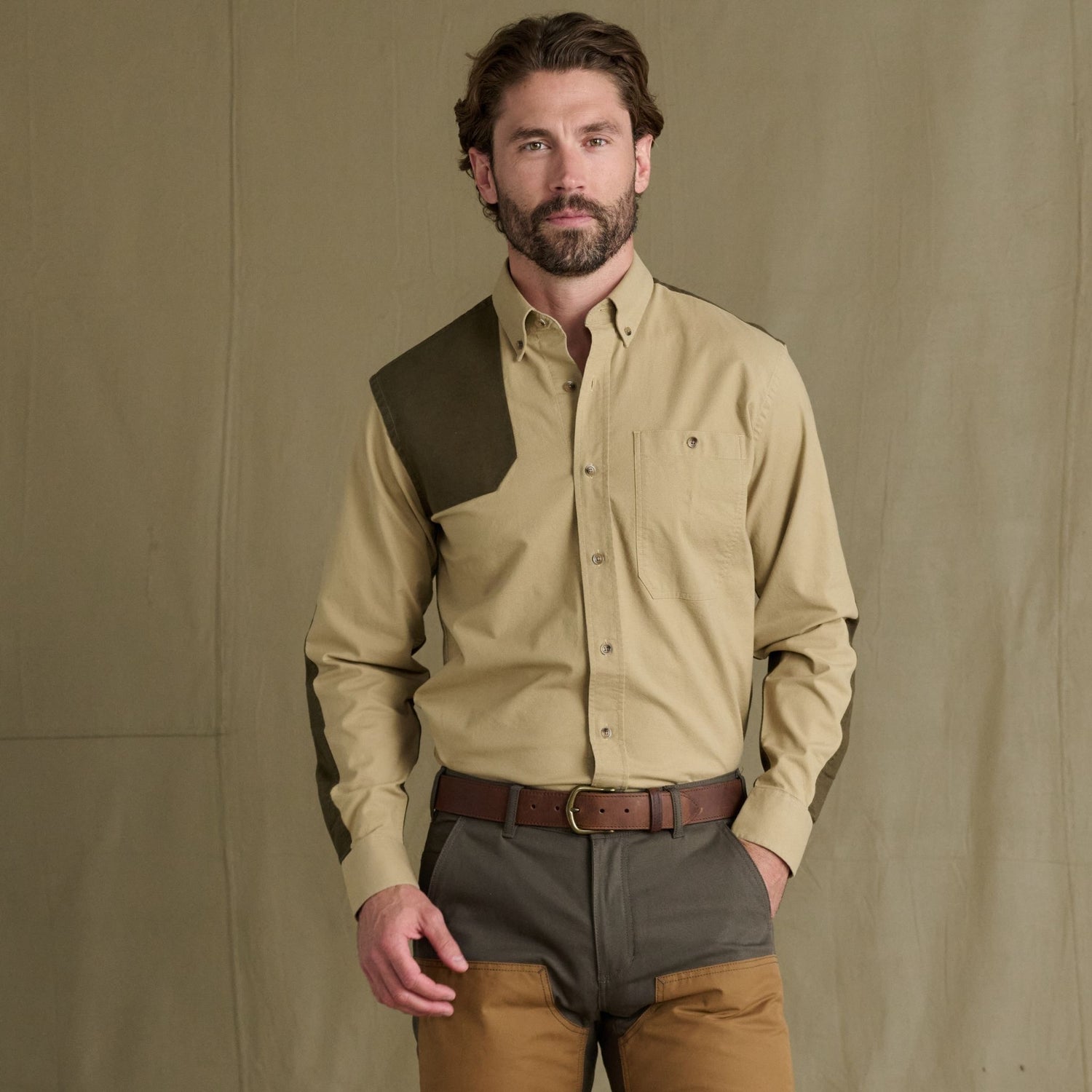 Man wearing Tom Beckbe Perry Field Shirt with green shoulder patch and brown pants, against a beige background.
