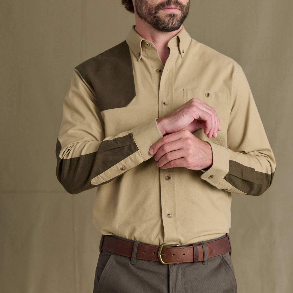Man wearing the Tom Beckbe Perry Field Shirt with olive patches and brown pants, adjusting his sleeve.