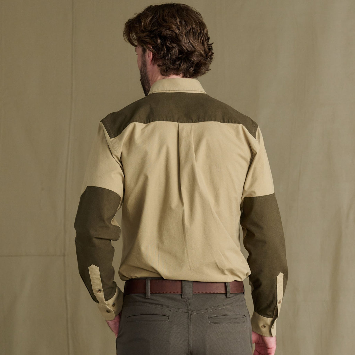 Man facing away, wearing a beige and olive Tom Beckbe Perry Field Shirt, gray trousers, against plain background.