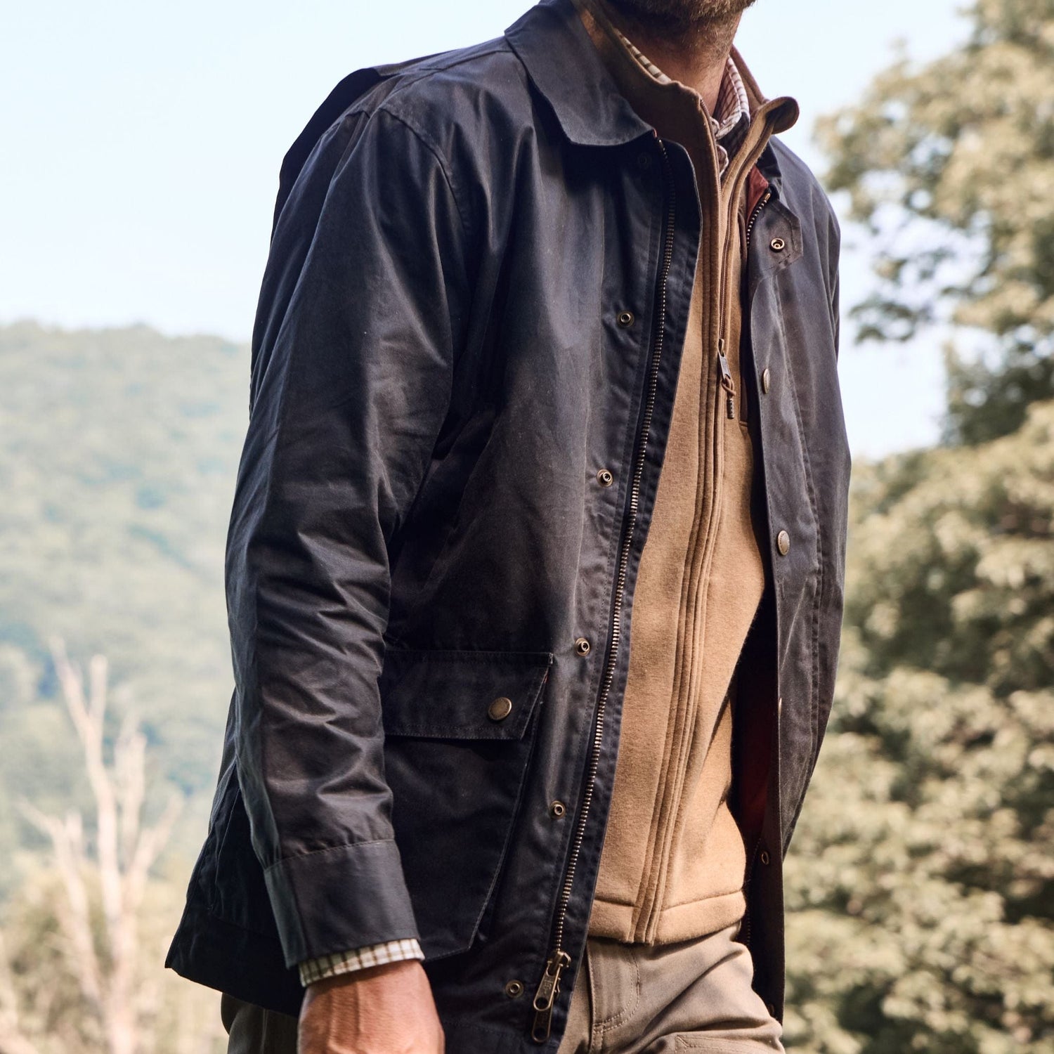 Man in a Tom Beckbe Piedmont Jacket over a beige sweater, strolling outdoors with trees and hills in the background.
