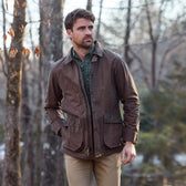 A man wears a Tom Beckbe Piedmont Jacket, green shirt, and tan pants while standing outdoors with hands in pockets.
