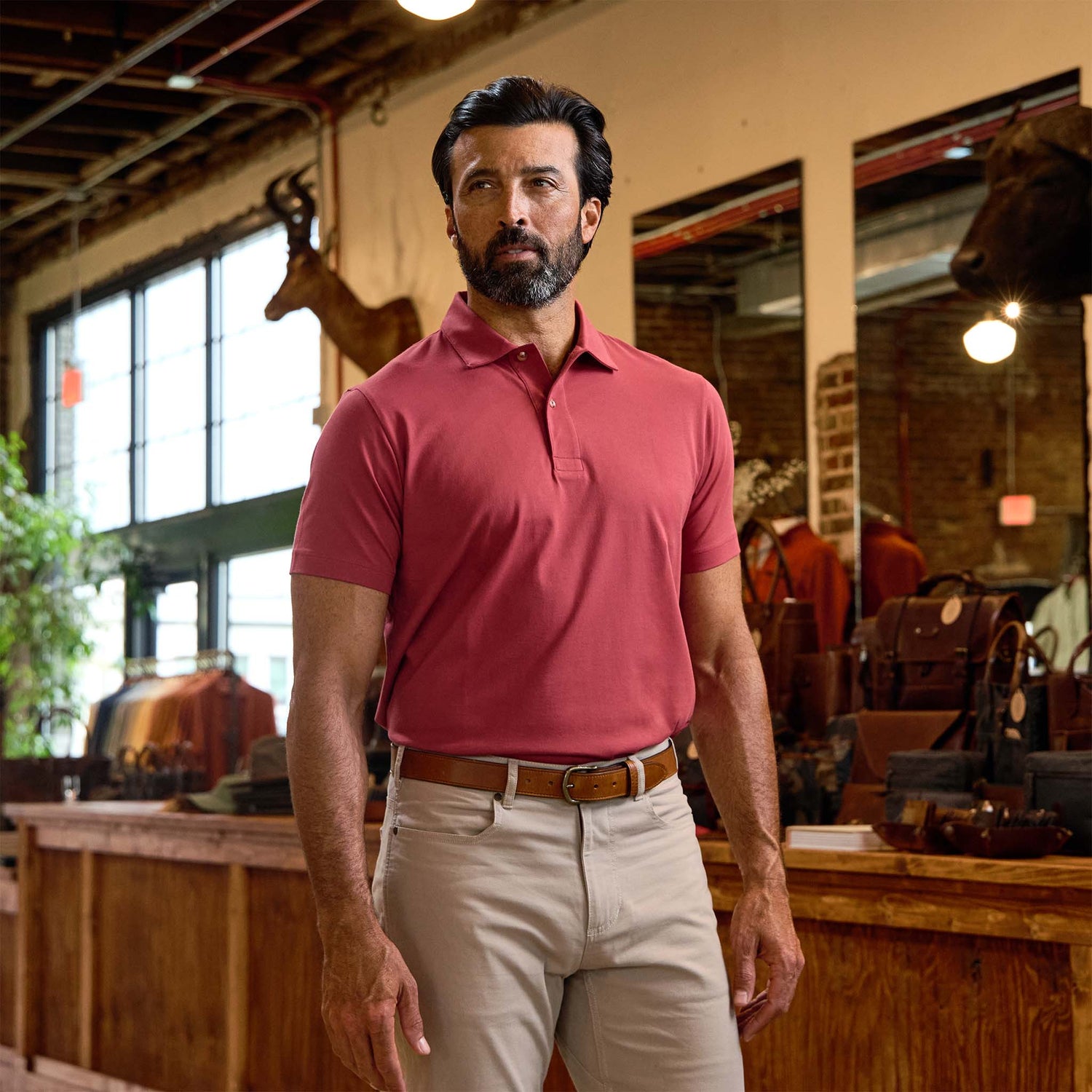 A man in a Tom Beckbe Benton Polo Shirt and beige pants stands in a store with leather goods and mounted animal heads.