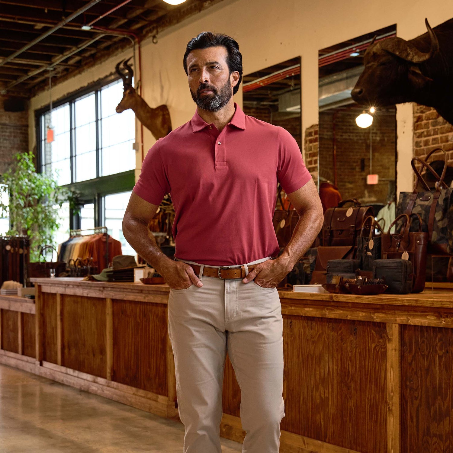Man in a Tom Beckbe Benton Polo Shirt and beige pants stands in a rustic store with leather bags and mounted animal heads.