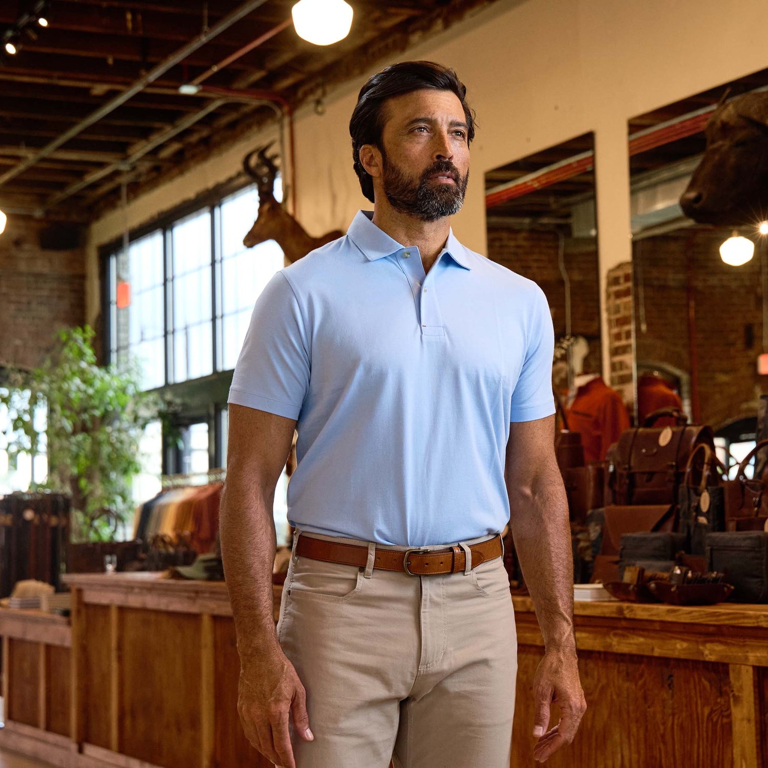 A man in a light blue polo shirt and beige pants stands indoors near wooden shelves and leather bags.