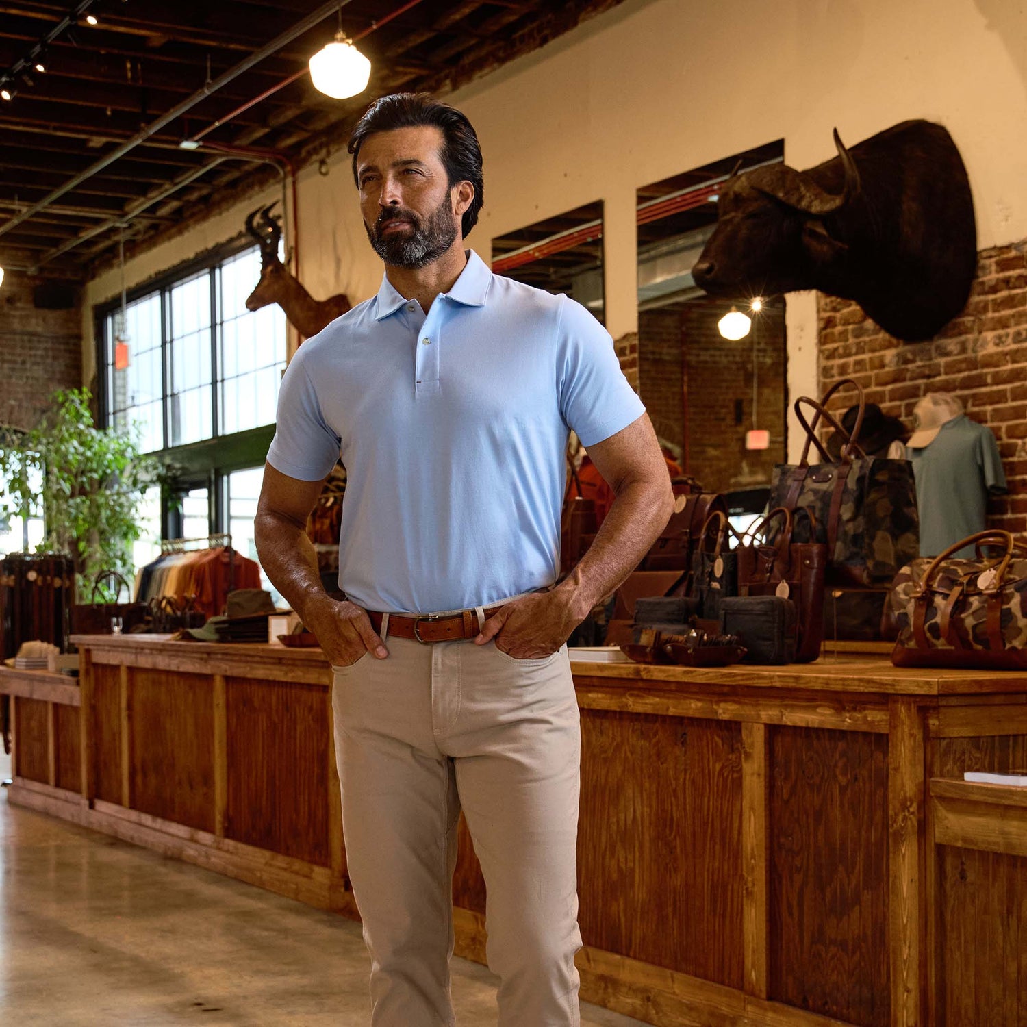 Man in a light blue polo shirt and beige pants stands in a rustic store with leather bags and animal mounts on the wall.