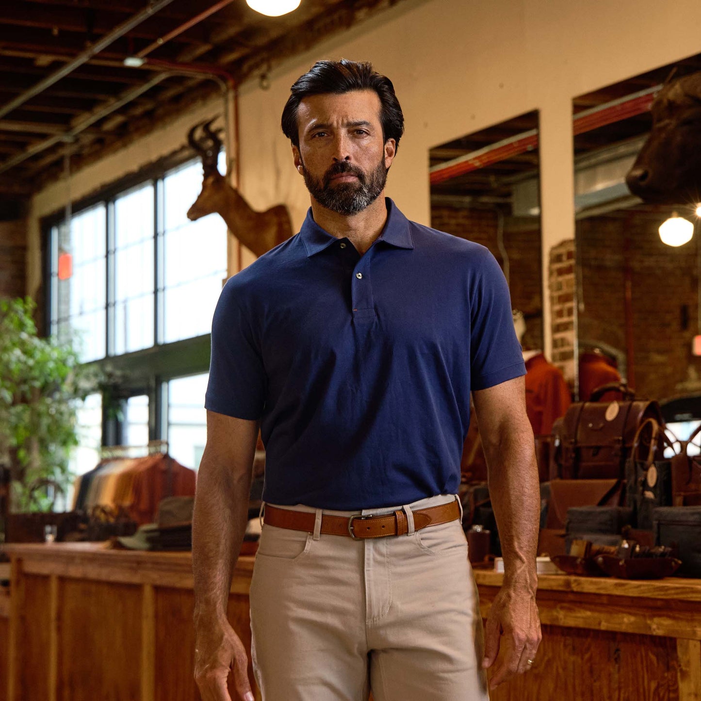 Man wearing a navy polo shirt and beige pants stands indoors in a rustic store with wood and leather decor.
