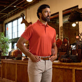 Man in orange polo shirt and beige pants stands indoors near wooden counter with bags and mounted animal heads in background.