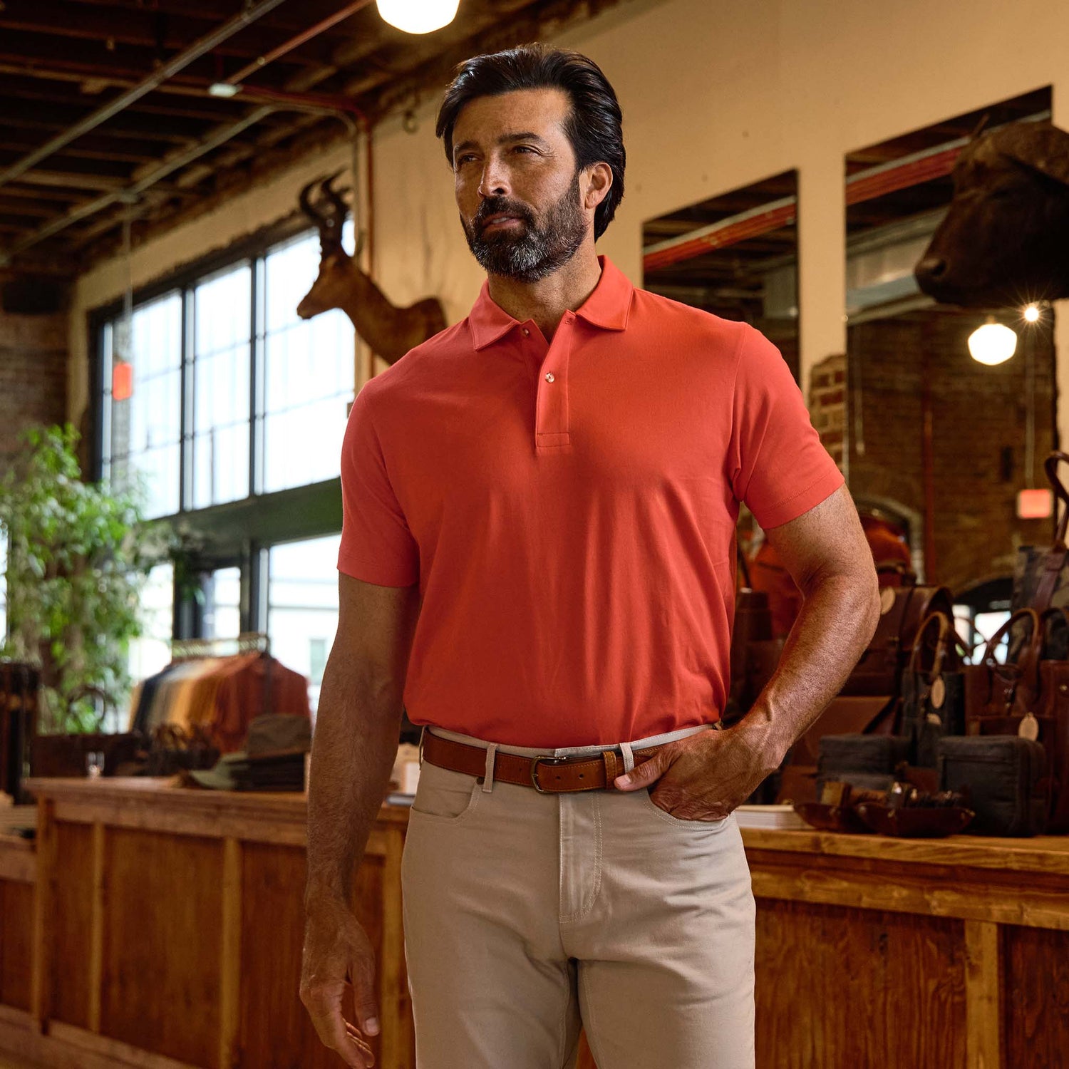 Man with dark hair and beard wears a Tom Beckbe Benton Polo Shirt and beige pants, standing indoors by a wooden counter.