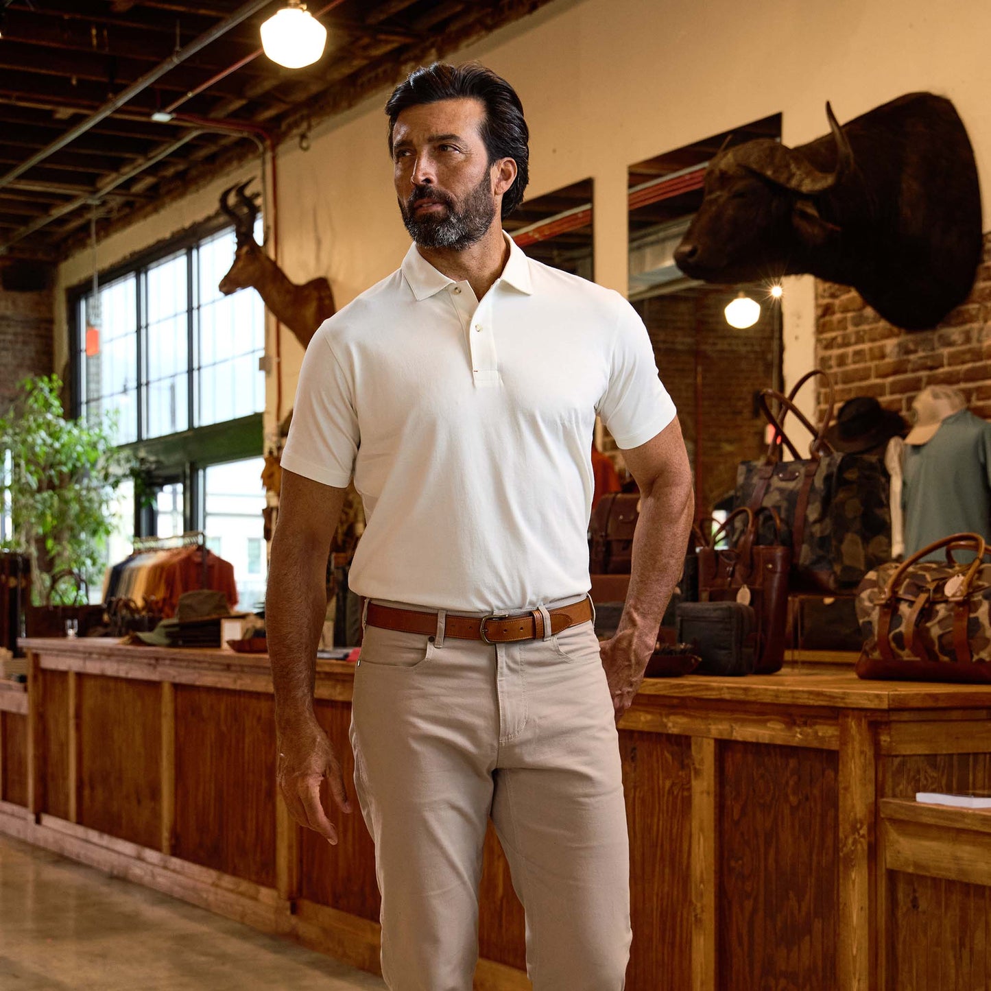 A man in a white polo shirt and beige pants stands in a rustic store with animal mounts and leather bags in the background.