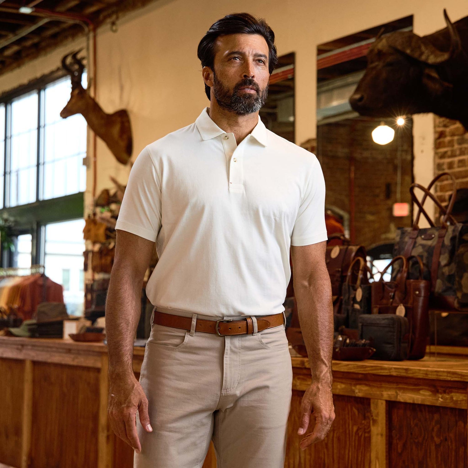 Man wearing a Tom Beckbe Benton Polo Shirt and beige pants stands in a rustic store with animal mounts and leather goods.