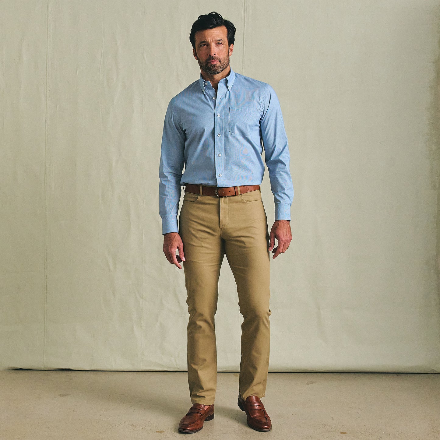 A man stands indoors wearing the Tom Beckbe Powell Performance Shirt, khaki pants, and brown dress shoes.