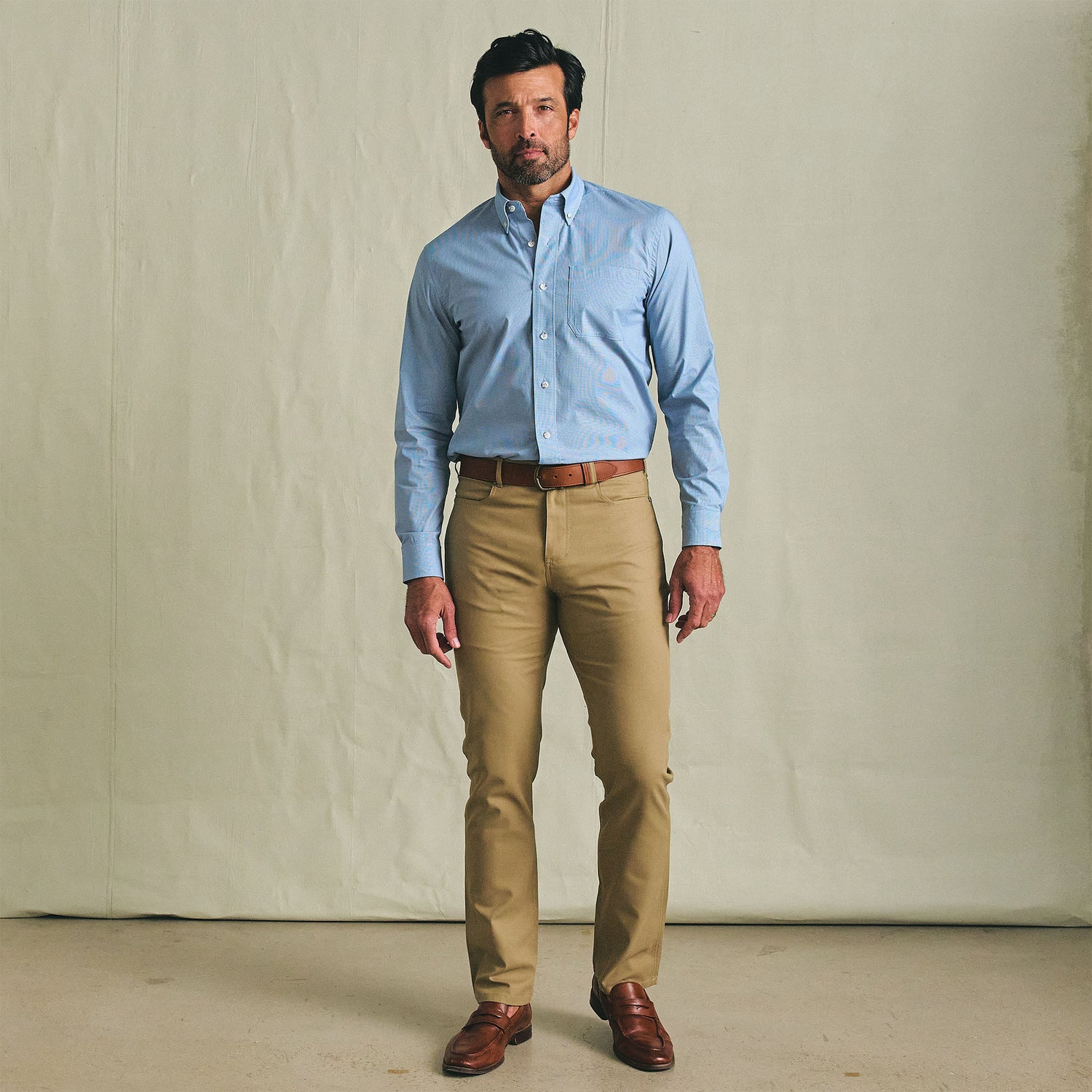A man stands indoors wearing the Tom Beckbe Powell Performance Shirt, khaki pants, and brown dress shoes.