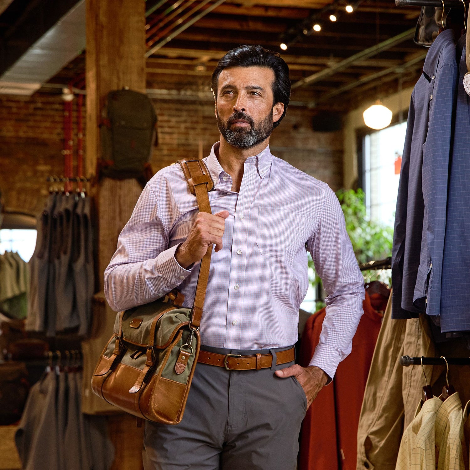 A man in a Tom Beckbe Powell Performance Shirt and gray pants carries a brown shoulder bag in a clothing store.