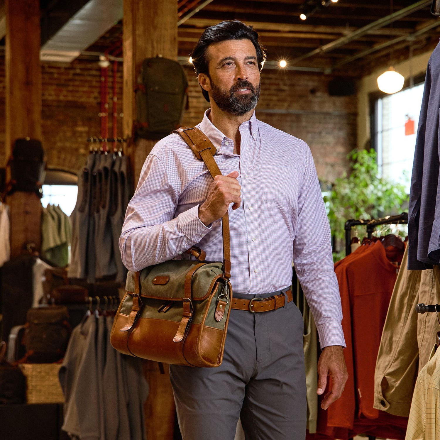 A man wears the Tom Beckbe Powell Performance Shirt and gray pants, carrying a brown leather bag in a clothing store.