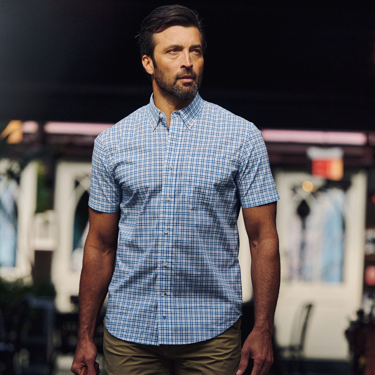 Man wears Tom Beckbe Powell Performance Shirt (Short Sleeve), blue plaid, and khaki pants outdoors with blurred background.