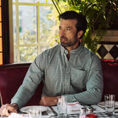 A man in a Tom Beckbe Powell Performance Shirt sits at a restaurant table with empty glasses, napkin, and plants behind him.