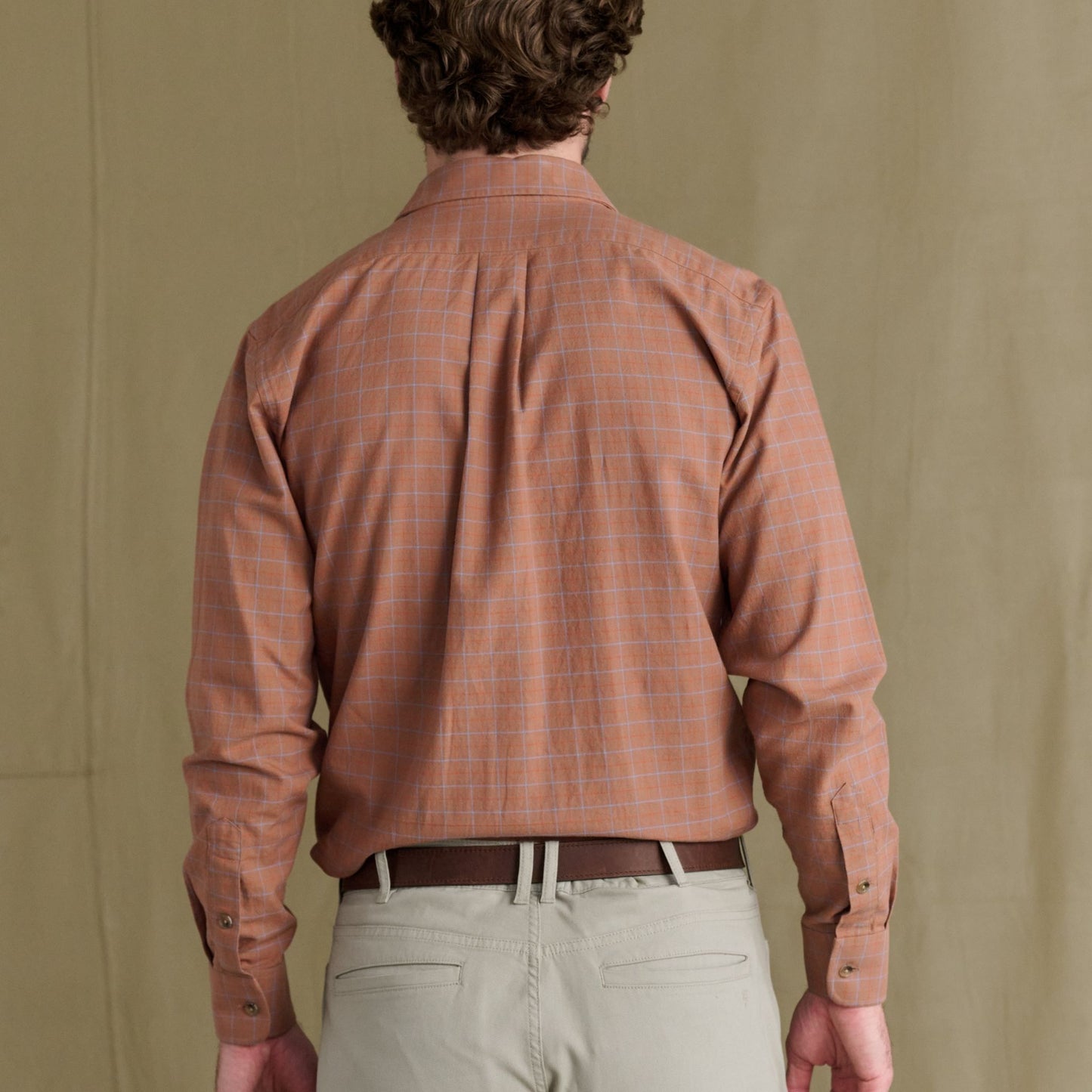 A person in a rust Prescott Estate Twill Shirt by Tom Beckbe and beige pants is shown from the back on a tan background.