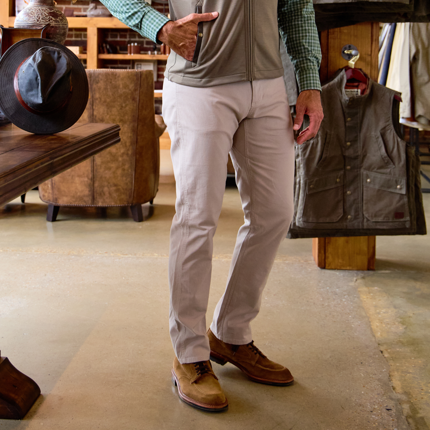 Man wears Tom Beckbe Reid Slim-Straight Leg Cotton Chino Pant, green checked shirt, and brown shoes indoors by a display.