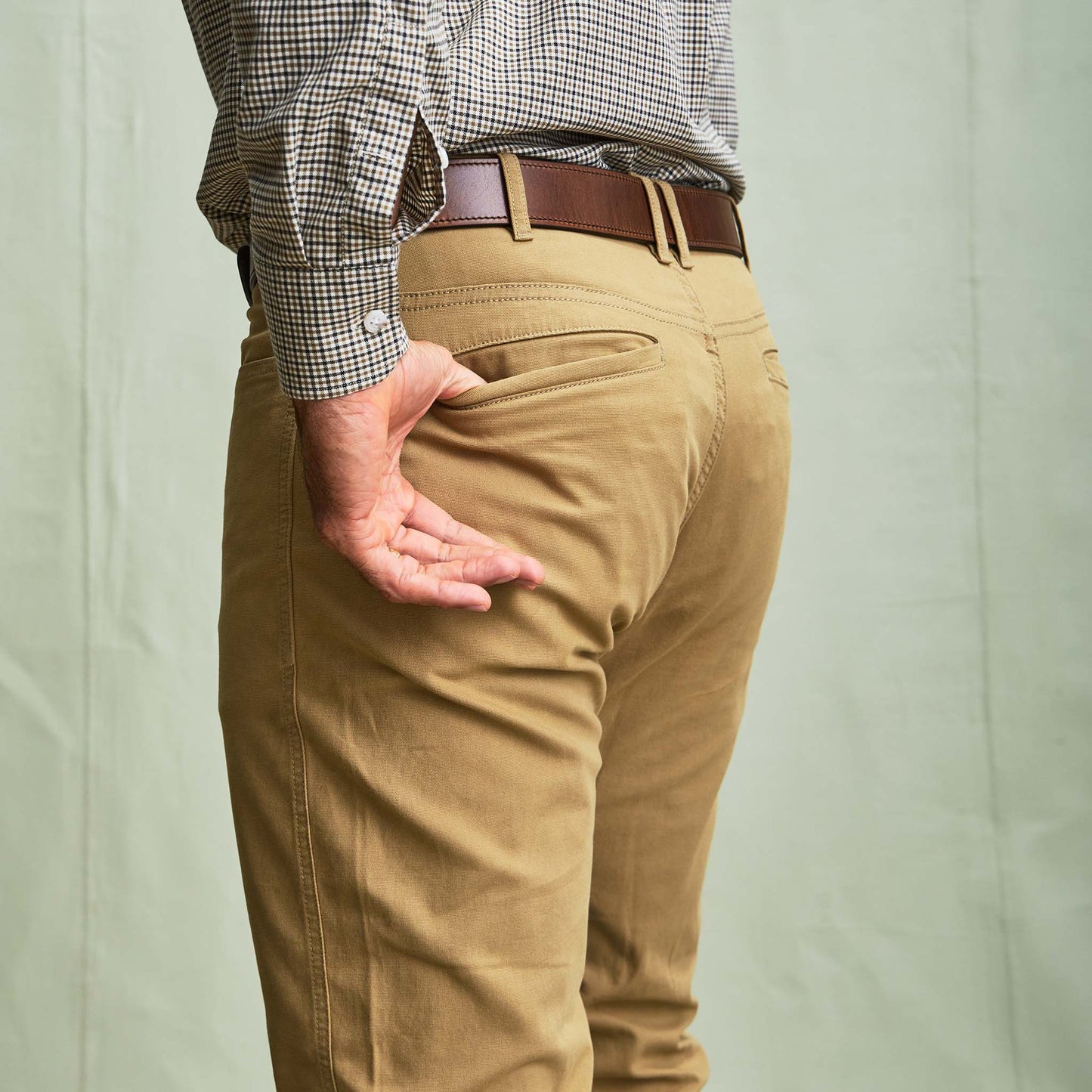 A person in Tom Beckbe's Reid Straight Leg Cotton Chino Pant and a brown belt stands with one hand in their back pocket.