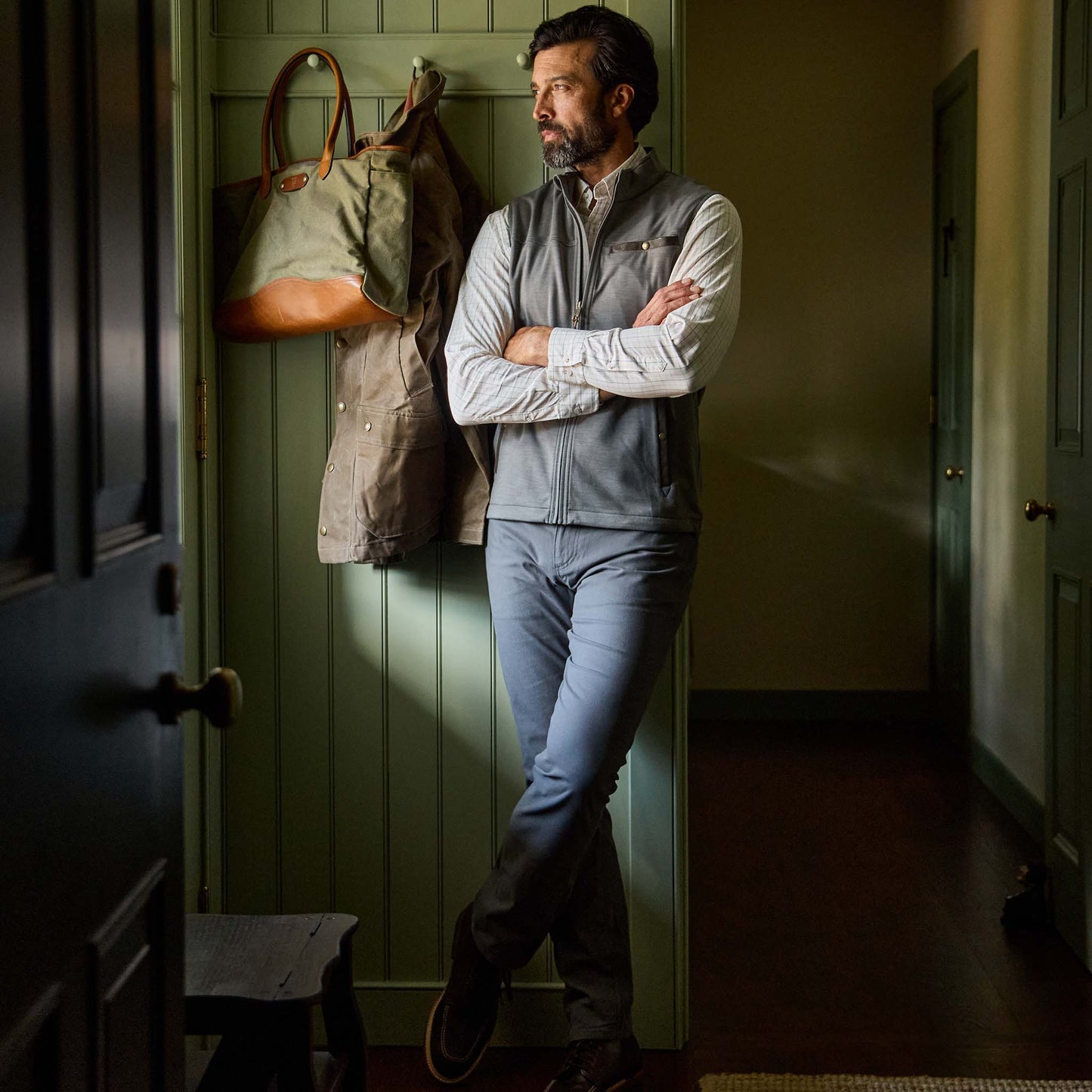 A man in Tom Beckbe's Reid Straight Leg Cotton Chino Pant stands with arms crossed against a green paneled wall.