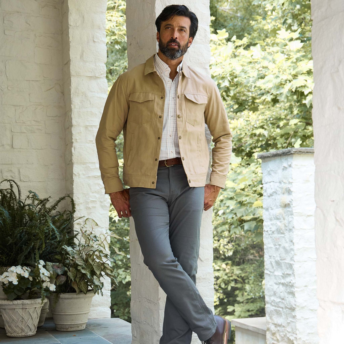 Man on a porch in a tan jacket, white shirt, and Tom Beckbe Reid Straight Leg Cotton Chino Pant; plants behind.