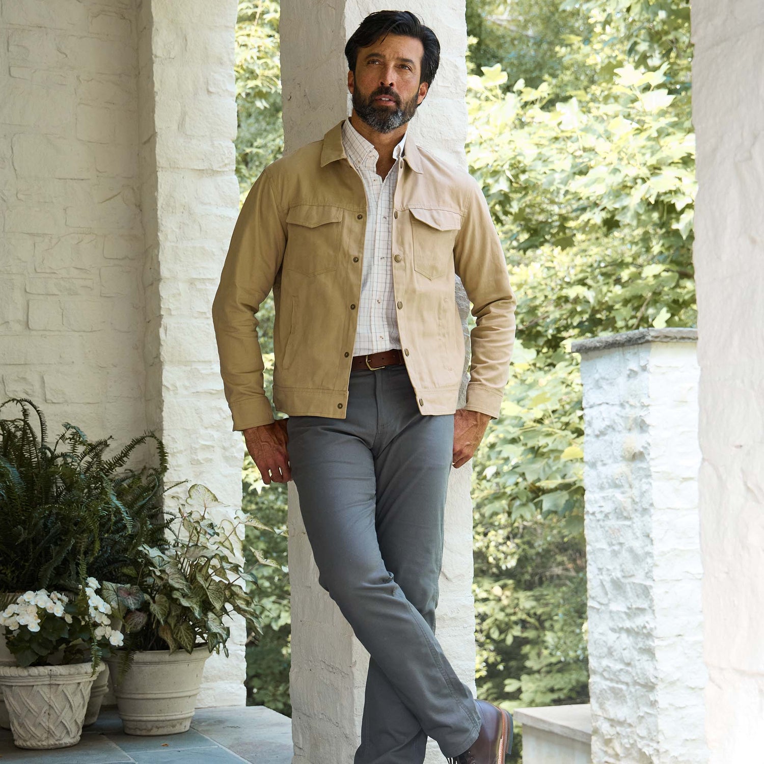 Man on a porch in a tan jacket, white shirt, and Tom Beckbe Reid Straight Leg Cotton Chino Pant; plants behind.