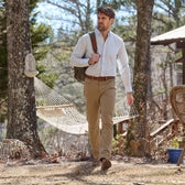 A man in Tom Beckbe Reid Straight Leg Cotton Chino Pants walks outdoors with a shoulder bag past a hammock and chair.
