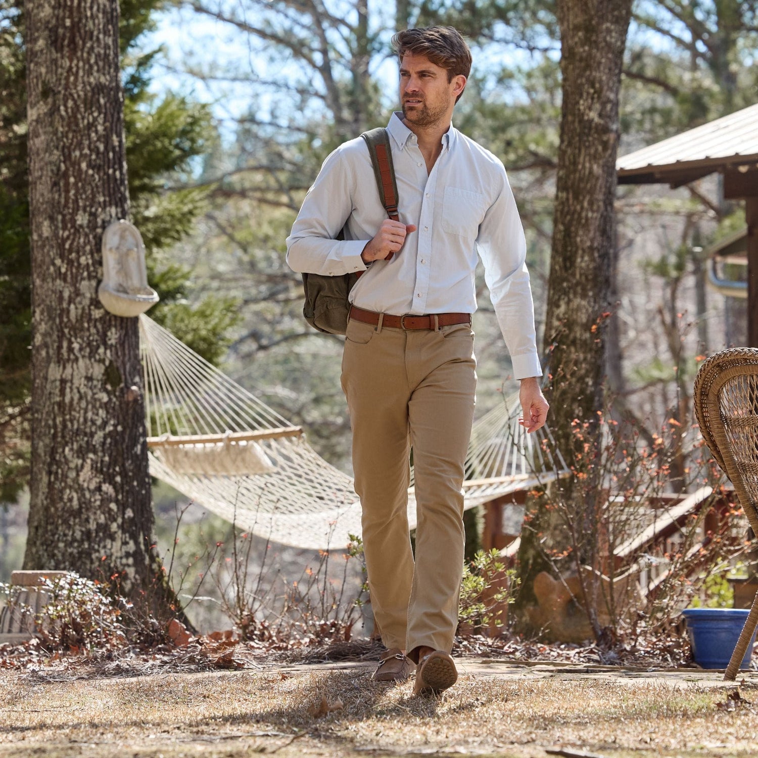 A man in Tom Beckbe Reid Straight Leg Cotton Chino Pants walks outdoors with a shoulder bag past a hammock and chair.