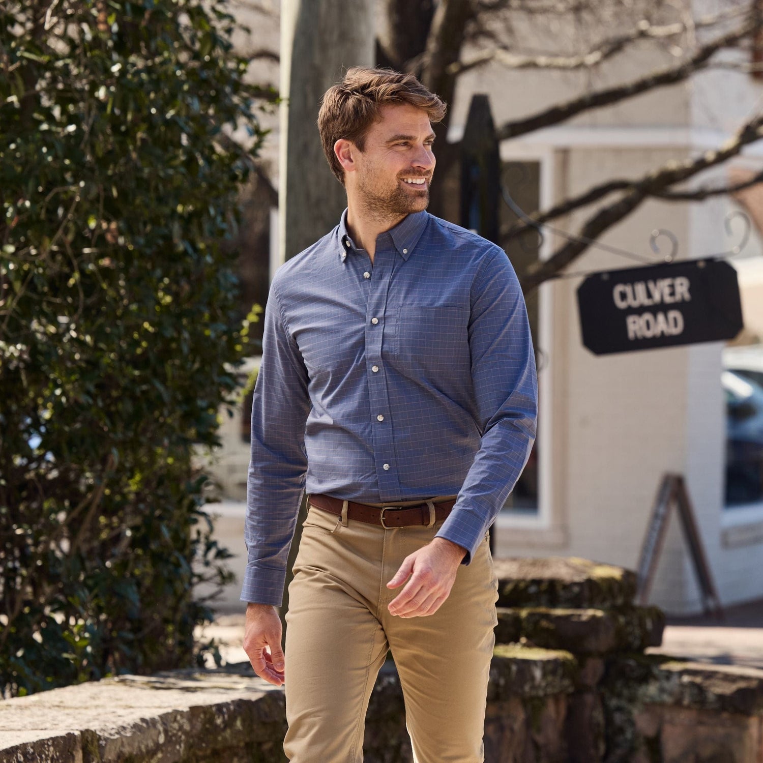 A man in a blue shirt and Tom Beckbe Reid Straight Leg Cotton Chino Pant walks outdoors by a stone wall and street sign.