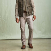 A man in Tom Beckbe Reid Straight Leg Cotton Chino Pants, brown boots, checked shirt, and gray vest stands by a plain background.