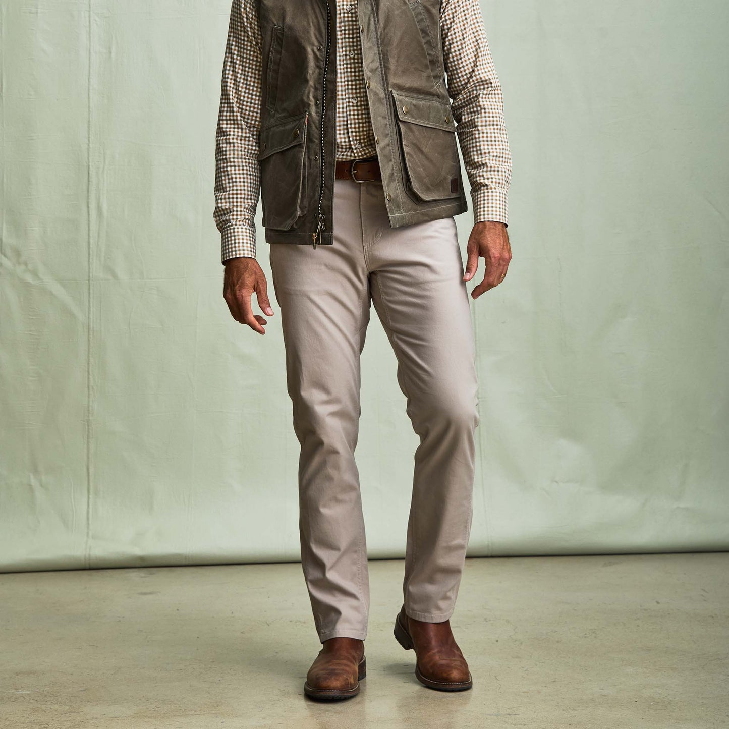 A man in Tom Beckbe Reid Straight Leg Cotton Chino Pants, brown boots, checked shirt, and gray vest stands by a plain background.