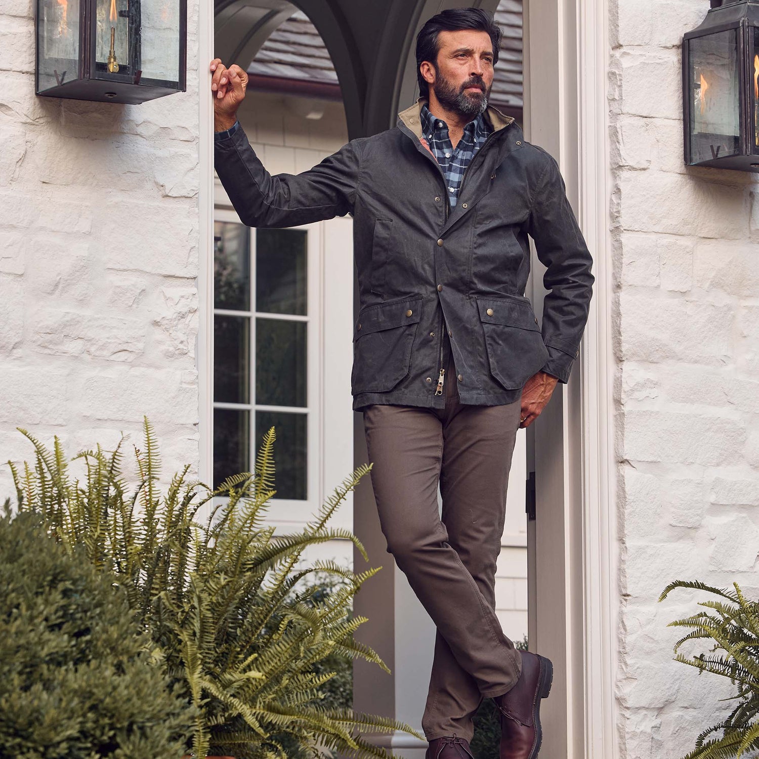 Man in a dark jacket and brown pants stands in a doorway, leaning against the frame with one leg crossed.