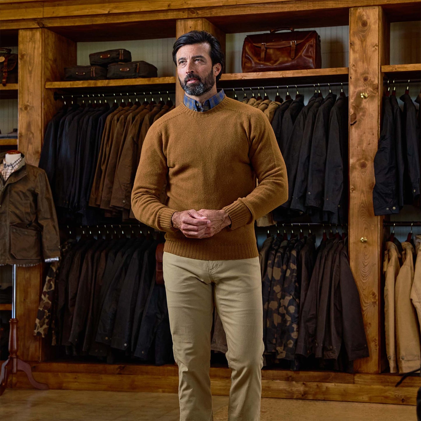 Man in a brown Tom Beckbe Saunders Crewneck Sweater and khaki pants stands before shelves of jackets and bags in-store.