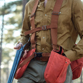 A person in a Tom Beckbe Strap Vest and brown shirt, holding a shotgun in the woods with orange pouches at their waist.