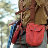 Person holding a shotgun, wearing Tom Beckbes brown strap vest in a forest setting.