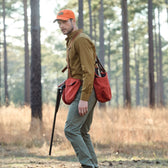 Wearing a Strap Vest and an orange cap, a hunter with a rifle walks through the forest, bag slung over their shoulder.
