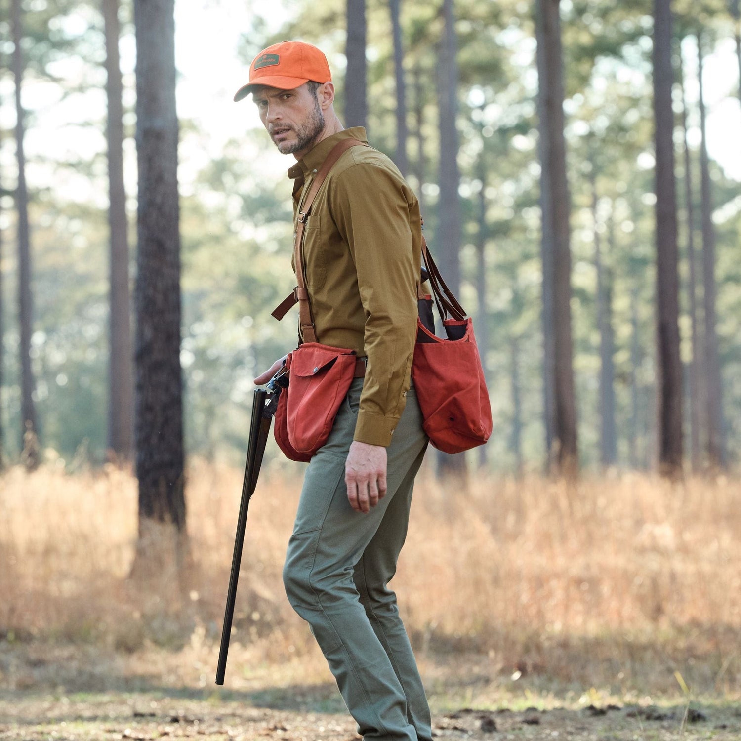 Wearing a Strap Vest and an orange cap, a hunter with a rifle walks through the forest, bag slung over their shoulder.