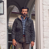 A man in a Tom Beckbe Tensaw Jacket, plaid shirt, and brown pants stands in a white brick doorway.