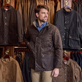 A man in a brown Tom Beckbe Tensaw Jacket stands in a store among racks of durable shelter cloth jackets.
