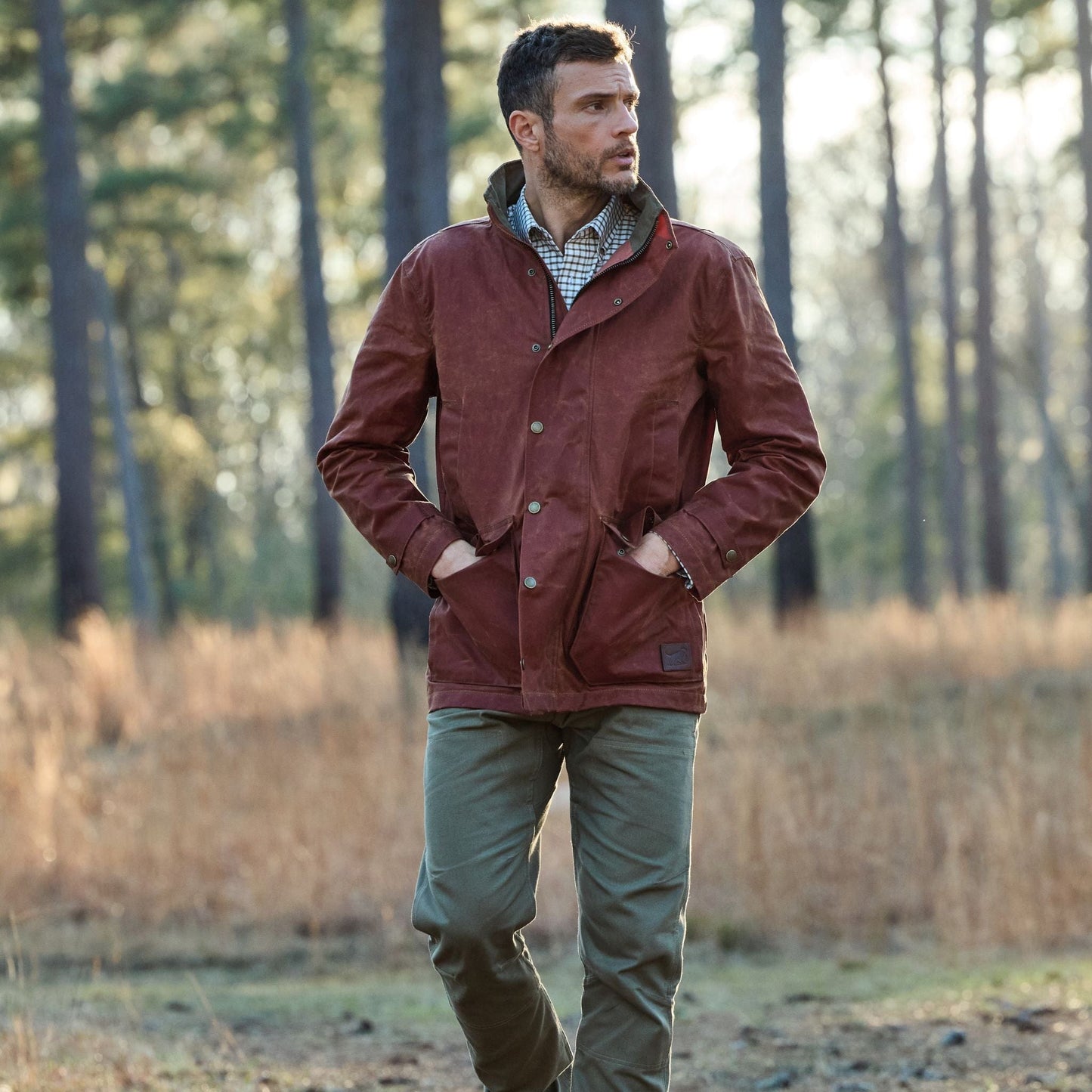 A man in a Fox Red Tensaw Jacket by Tom Beckbe and green pants walks in a forest with sunlight filtering through the tall trees.